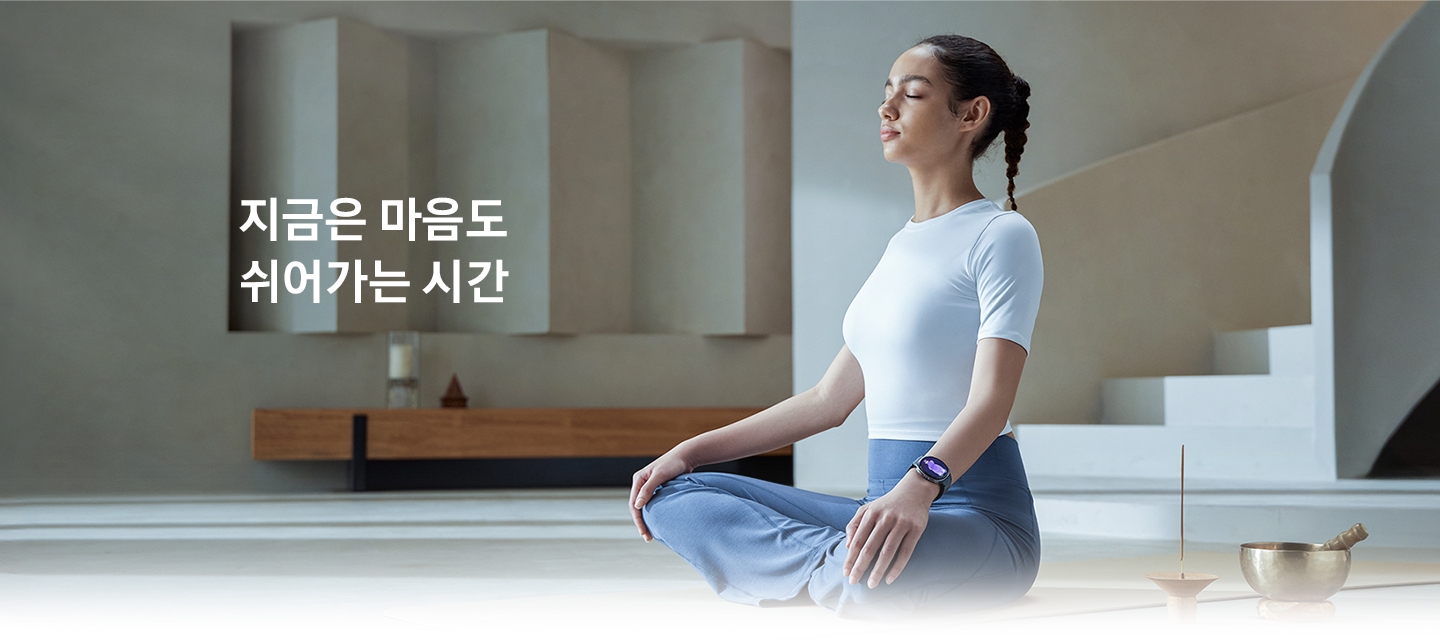 한 여성이 실내에 가부좌 자세로 앉아 눈을 감고 있습니다. 손목에는 갤럭시 워치8을 착용하고 있습니다.