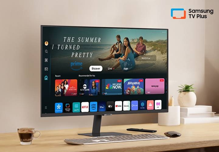 책상 위에 모니터가 있습니다. 화면 안에는 다양한 OTT 앱과 연결할 수 있는 홈 UI가 띄워져 있습니다. 오른쪽 상단에는 Samsung TV Plus 로고가 있습니다.