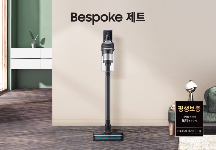 모던한 분위기의 거실이 보입니다. 왼쪽에는 해가 들어오는 넓은 창이 보이고 그 옆에는 하얀색 선반형 벽 수납장이 보입니다. 수납장 위에는 초록 잎 2장이 자라고 있는 작은 화분이 올려져 있습니다. 선반 뒤 쪽 벽은 부드럽고 잔잔한 초록색 톤을 띄고 있습니다. 마루는 헤링본 스타일의 진한 월넛 색상입니다. 정면에 보이는 넓은 벽면은 진한 아이보리 톤을 띄고 있습니다. 오른쪽 구석으로는 진한 그린 색의 패브릭 소파가 있습니다. 소파 위에는 아이보리 톤의 사각 쿠션과 굵은 짜임의 니트 재질 담요가 흘러내리 듯 올려져 있습니다. 소파 앞에는 우드 재질의 원형 테이블이 있고, 바닥엔 크림색의 러그가 깔려 있습니다. 소파 왼쪽 끝부분엔 우드 패널 위로 책 두 권과 금색 빛의 오브 재가 있습니다. 거실 전경 가운데에는 Bespoke 제트 문구가 크게 적여있고 문구 왼쪽으로 Bespoke 제트 210W 정면을 바라보고 서 있습니다.