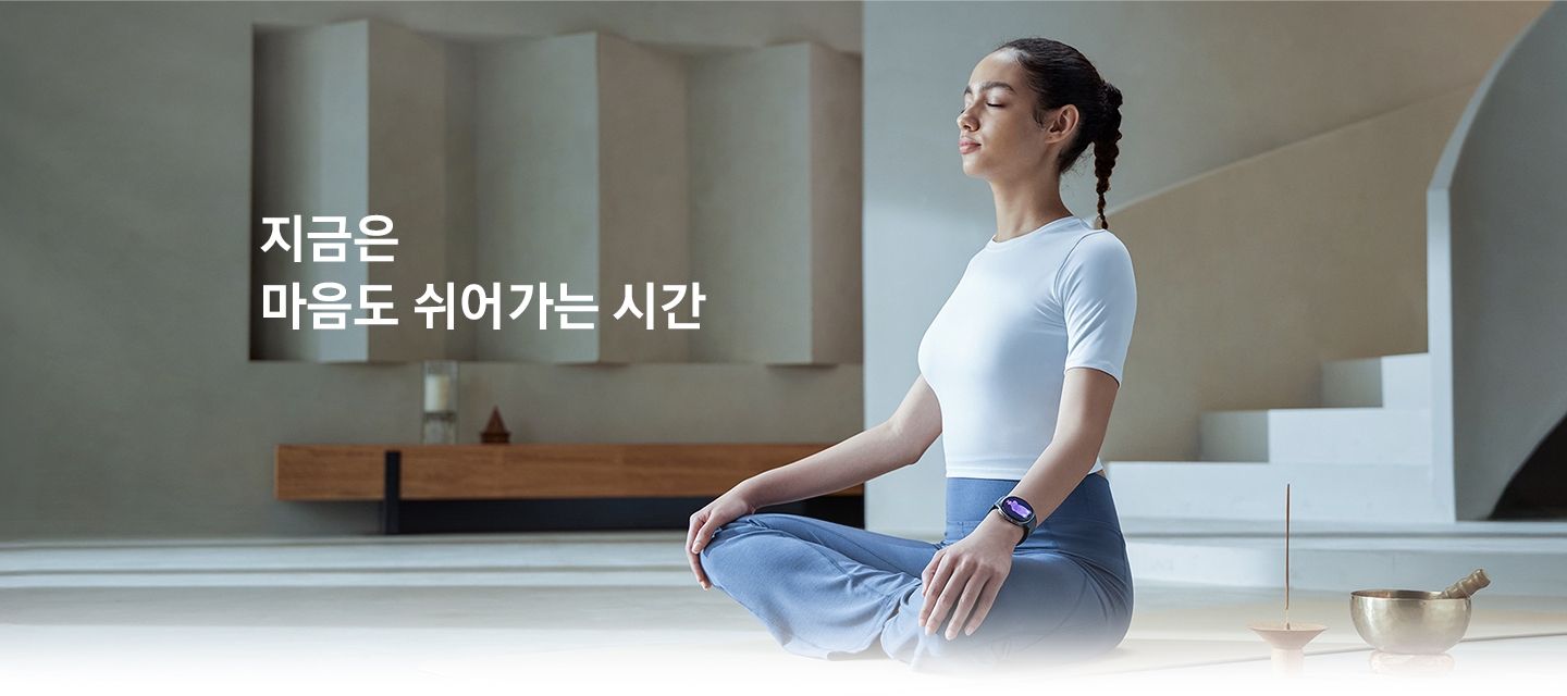 한 여성이 실내에 가부좌 자세로 앉아 눈을 감고 있습니다. 손목에는 갤럭시 워치8을 착용하고 있습니다.