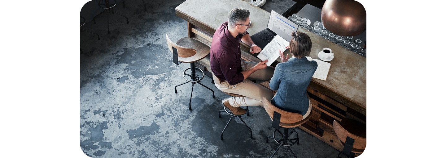 Two people sitting at the bar of a coffee shop, looking at papers and a laptop