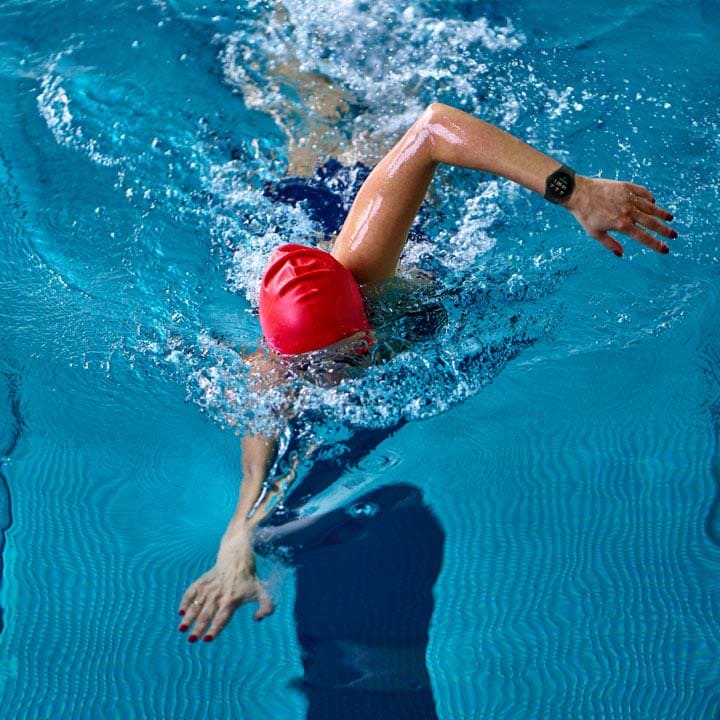ไอคอนการออกกำลังกายแบบต่าง ๆ และมีไอคอนของการว่ายน้ำถูกไฮไลท์อยู่ ผู้หญิงคนหนึ่งว่ายน้ำอยู่ในสระว่ายน้ำขณะที่สวม Galaxy Watch4 อยู่