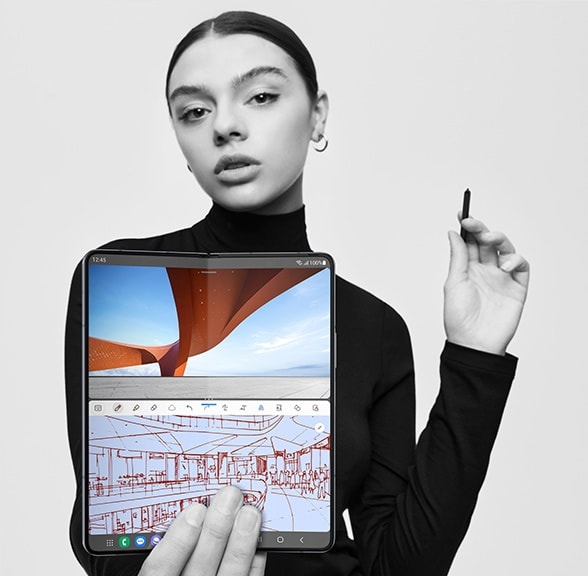 A woman holds an open Galaxy Z Fold5 with the Main Screen facing away from her and toward the viewer. She holds S Pen Fold Edition for Galaxy Z Fold5 in her hand. On the top half of the screen is a vibrant photo of modern architecture. On the lower half is an architectural sketch in a note-taking app.