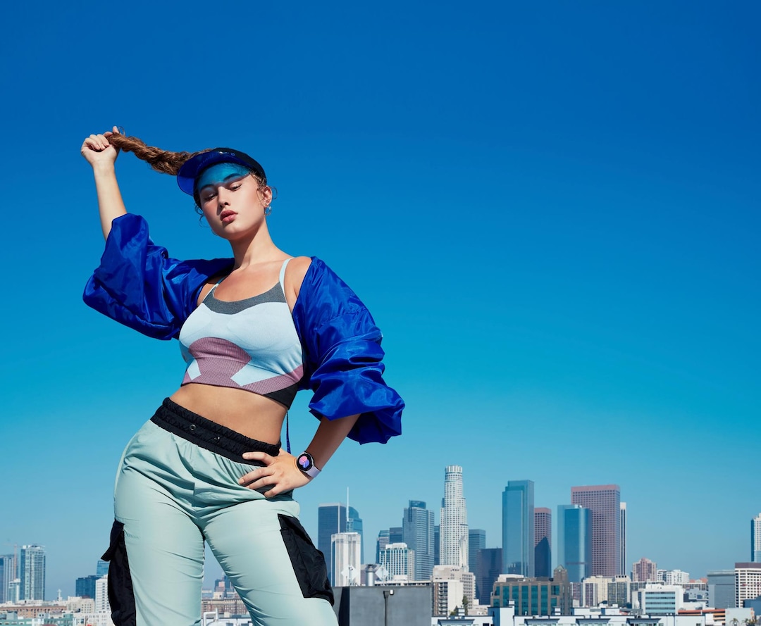 A woman striking a pose outdoors in front of the city skyline wearing a blue outfit with a Galaxy Watch5 on her wrist showing the time as '5' in a blue and pink gradient.