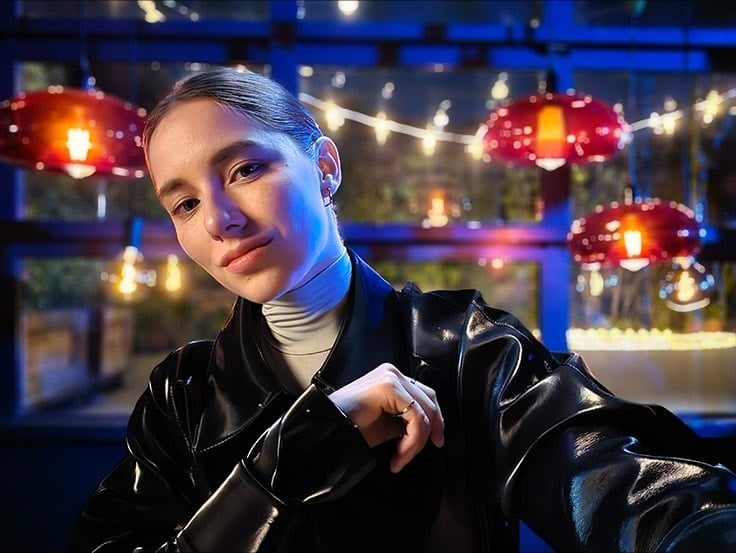 A woman takes a colorful, clear selfie in a dark setting with string lights and lanterns behind her. The photo was taken with Galaxy S23 Ultra using wide mode with an aperture of F2.2. 