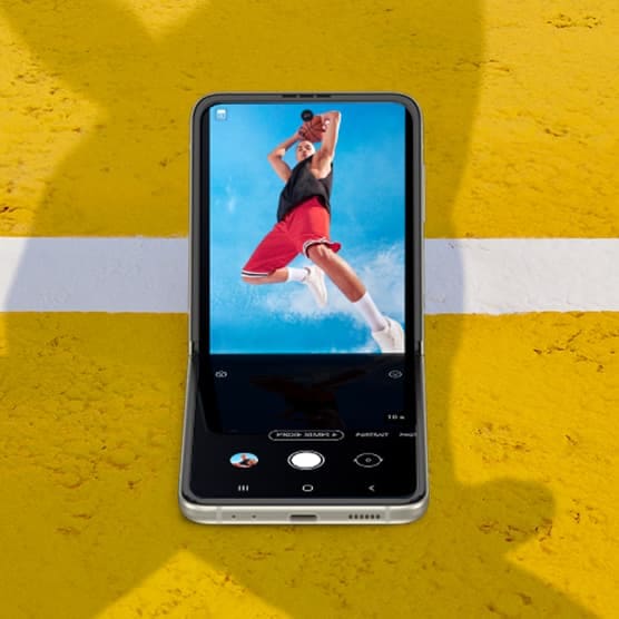 Un homme tenant un ballon de basketball au-dessus de sa tête et sautant en regardant vers la caméra. Le Galaxy Z Flip3 5G vu en mode Flex avec l’application Appareil photo sur l’écran principal et la même photo vue dans le viseur.