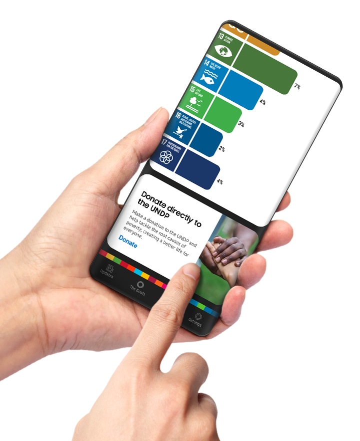 손에 들린 스마트폰 화면에 삼성 글로벌 골스 앱 페이지가 보입니다. 검지로 목표에 대한 직접 기부를 안내하는 카드를 터치하고 있습니다. 사용자는 광고를 보고 적립금을 모아 기부할 수도 있지만, 원하는 목표에 직접 기부할 수도 있습니다.