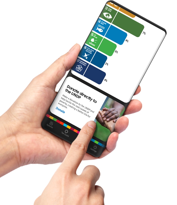 손에 들린 스마트폰 화면에 삼성 글로벌 골스 앱 페이지가 보입니다. 검지로 목표에 대한 직접 기부를 안내하는 카드를 터치하고 있습니다. 사용자는 광고를 보고 적립금을 모아 기부할 수도 있지만, 원하는 목표에 직접 기부할 수도 있습니다.