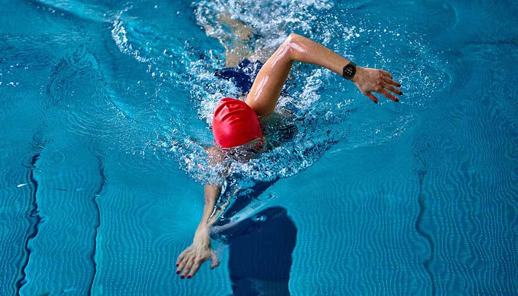 Se pueden ver iconos de diferentes ejercicios y el icono de natación está resaltado. Una mujer nada en una piscina mientras usa un dispositivo Galaxy Watch4.