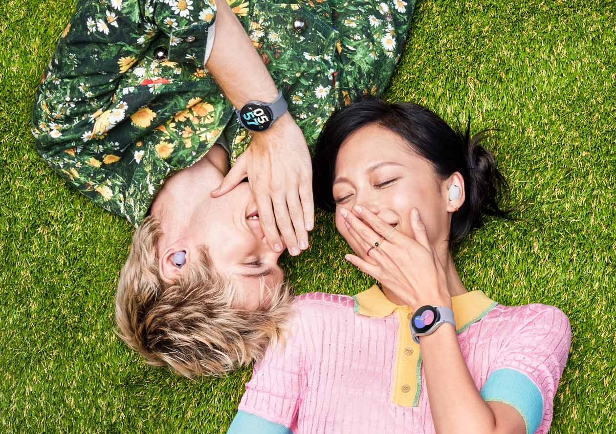 Two people lying next to each other on the ground. The guy on the left positioned upside down is facing the girl on the right, while the girl on the right is left side up with both their heads next to each other. Both are joyfully having a conversation with a Galaxy Buds2 Pro in their ear. Both people also have a Galaxy Watch5 on their wrists.