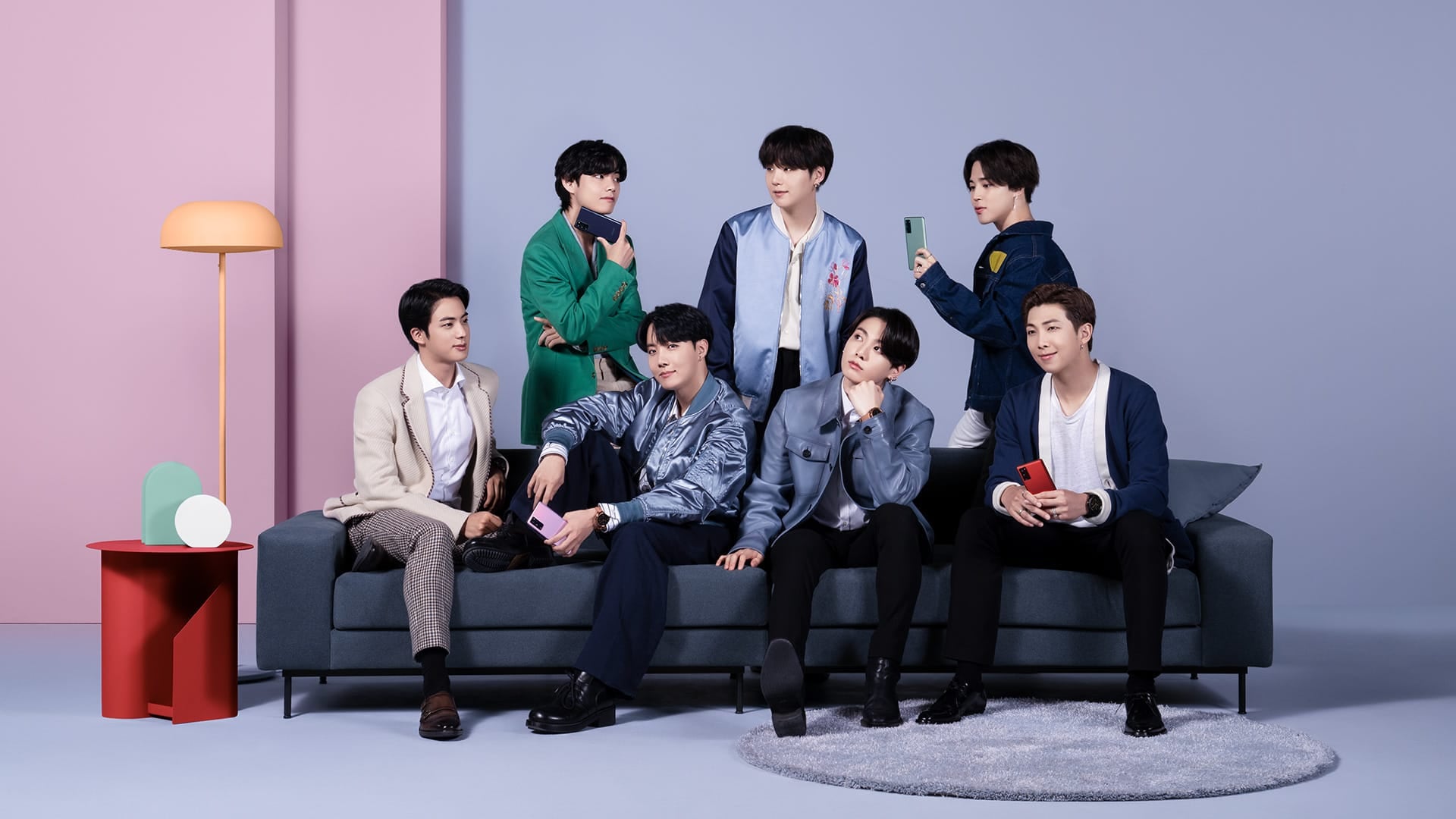 Les membres de BTS, quatre assis sur un canapé et trois debout a l’arriere. Ils portent chacun les différentes couleurs du téléphone Galaxy S20 FE.