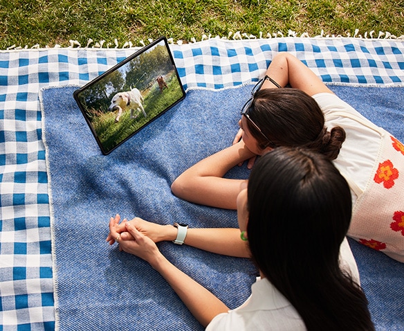 Dos mujeres recostadas sobre una manta de picnic en el césped mientras miran un video en un dispositivo de la serie Galaxy Tab  S9 con la Book Cover inteligente puesta en modo horizontal, apoyada utilizando la cubierta posterior plegable.