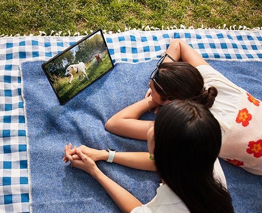 Dos mujeres recostadas sobre una manta de picnic en el césped mientras miran un video en un dispositivo de la serie Galaxy Tab  S9 con la Book Cover inteligente puesta en modo horizontal, apoyada utilizando la cubierta posterior plegable.