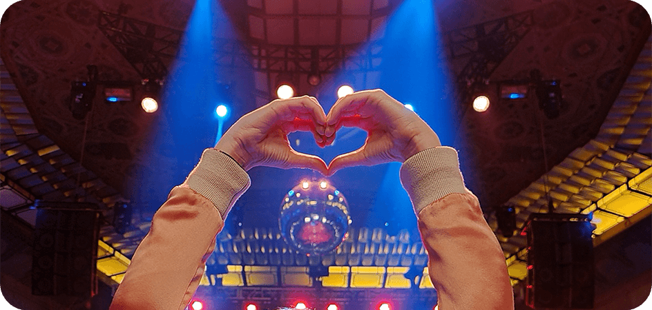 Foto tomada con el modo noche del Galaxy S20 Ultra en un lugar oscuro en un concierto con una mujer en el medio haciendo un corazón con las manos. La foto está bien iluminada y es clara a pesar del ambiente con poca luz