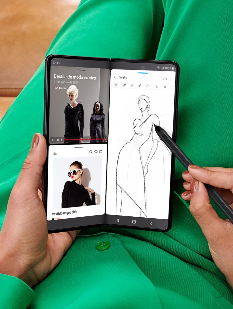 Unas manos sosteniendo un Galaxy Z Fold3  desplegado y dibujando con el S Pen Fold Edition. La pantalla principal está en modo Multi Active Window. La mitad de la pantalla muestra dos instancias de la aplicación de Internet abierta, una con una transmisión en vivo de un desfile de modas y la otra con un sitio web que muestra imágenes de las prendas del desfile. La otra mitad de la pantalla es un boceto de un vestido que se está creando en Samsung Notes.