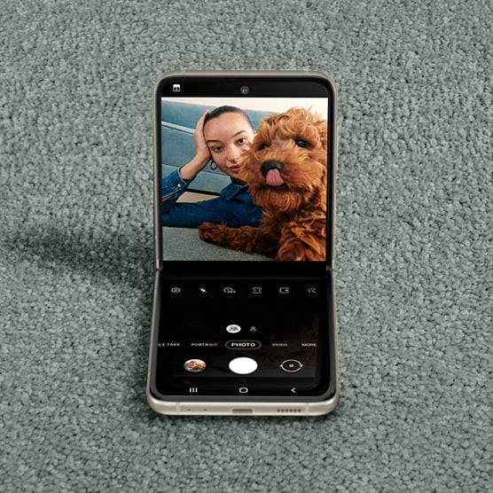 Eine Frau macht ein Selfie mit einem Welpen, der sich über die Nase leckt. Das Galaxy Z Flip3 5G ist im Flex-Modus mit der Kamera-App auf dem Hauptdisplay und demselben Foto im Sucher zu sehen.