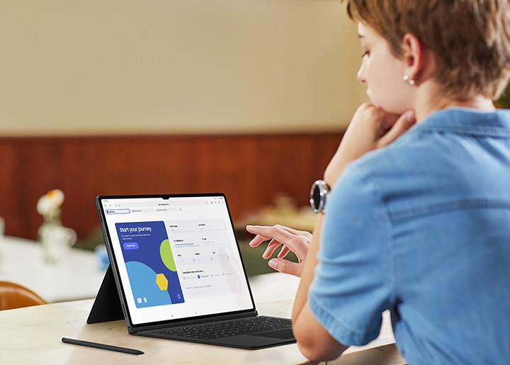 A woman at a cafe is working on a brochure using a Galaxy Tab S9 Series device with Book Cover Keyboard on. An S Pen is placed next to the tablet.
