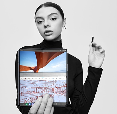 Une femme tient un Galaxy Z Fold5 ouvert, l’écran principal tourné vers l’objectif. Elle tient le S Pen Fold Edition pour Galaxy Z Fold5 dans sa main. Sur la moitié supérieure de l’écran se trouve une photo éclatante d’architecture moderne. Sur la moitié inférieure on voit un croquis architectural dans une application de prise de notes.