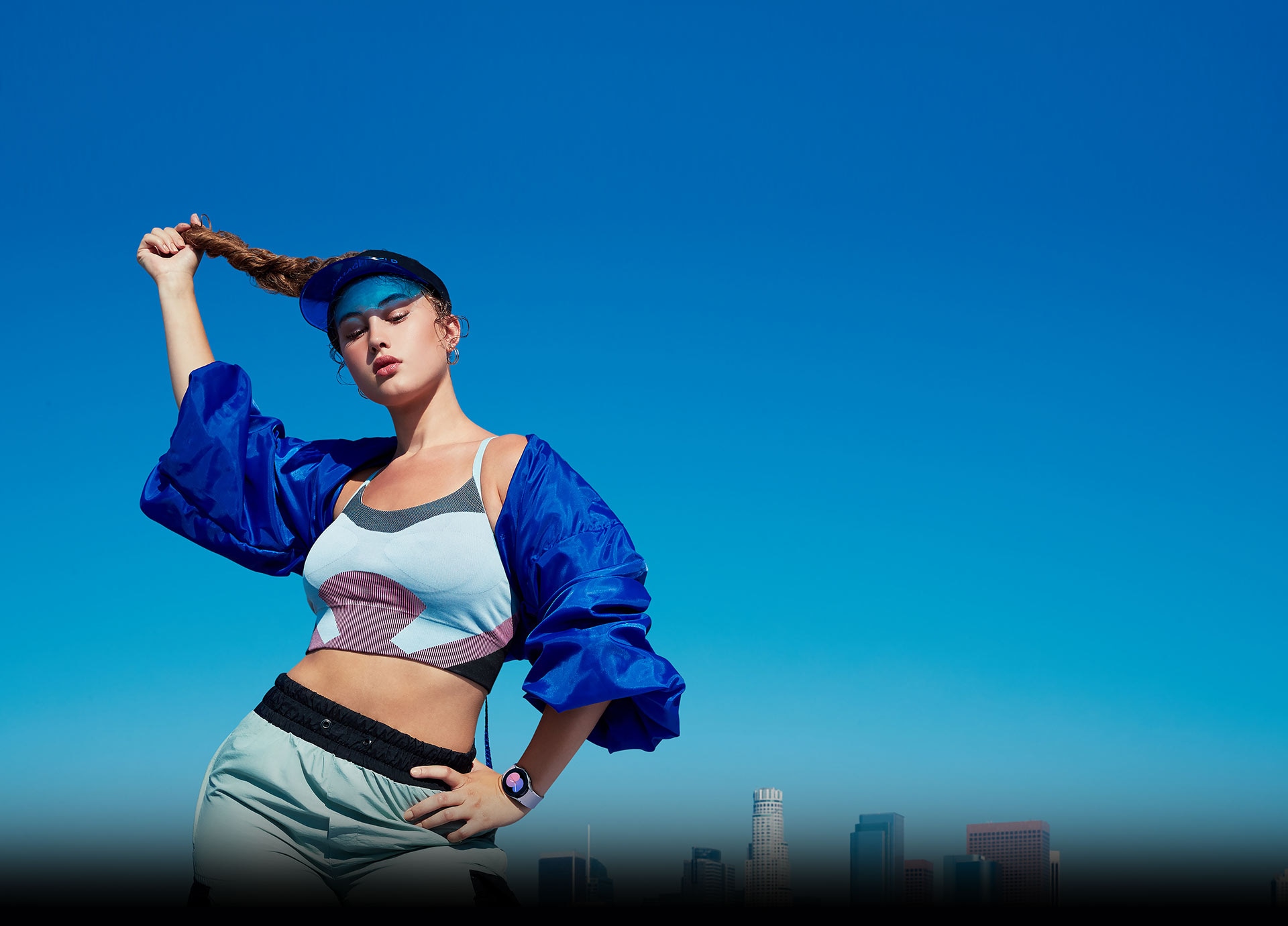 A woman striking a pose outdoors in front of the city skyline wearing a blue outfit with a Galaxy Watch5 on her wrist showing the time as '5' in a blue and pink gradient.