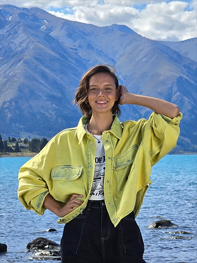 Ein mit 6,5-fachem Zoom aufgenommenes Foto einer Frau, die an einem See posiert, ist lebendig und die Berglandschaft im Hintergrund ist scharf.