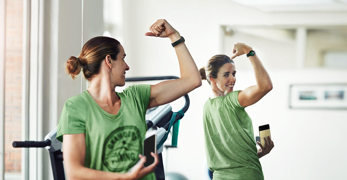 A woman is looking at the mirror and flexing her muscles while wearing a Galaxy Watch4 device and holding a Galaxy Z Flip3 5G in her right hand.