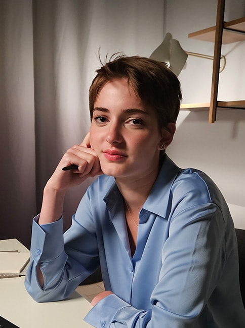 A woman is seated at a table in a low-lit room. The textures around the room are detailed and the colors of her clothes and skin tone are natural and vibrant.