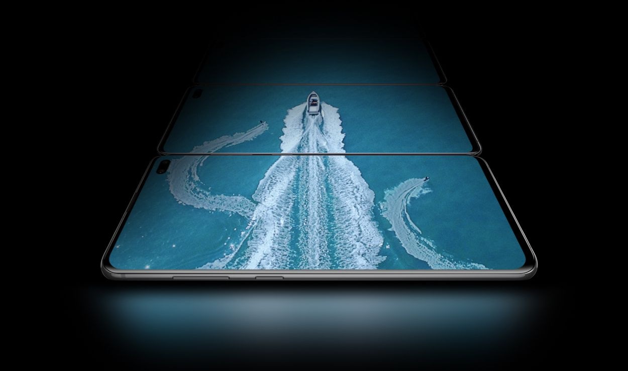 Three Galaxy S10 plus phones in landscape mode, laying flat above the one in front of it, seen at a slight angle from the Bixby and volume button side. On the three screens there is an aerial shot of a boat in water with two water skiers, showing the expansiveness of the Infinity-O Display.