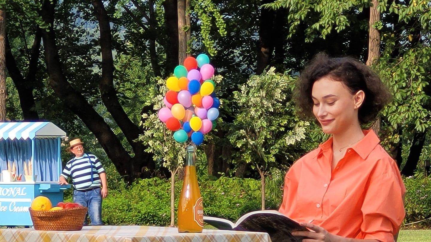 10x zoomed in photo, getting even closer to the woman sitting alone at a table. More details become clear, like that she's reading a magazine and on top of the table is a bunch of many mini balloons and an orange bottle.
