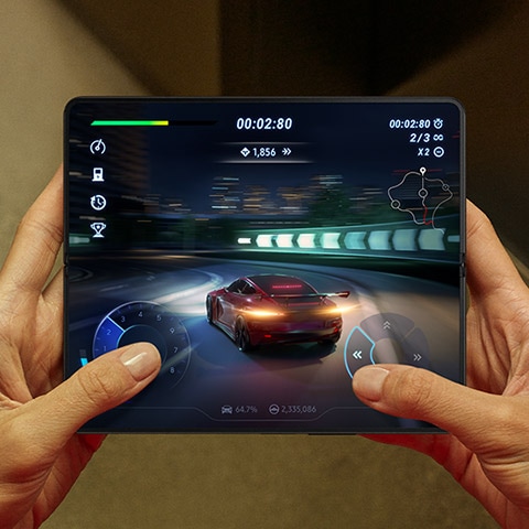 Hands holding an unfolded Galaxy Z Fold4. The Main Screen displays a scene from a racing game. A sports car speeds down a highway with a dashboard overlay on the screen. The content is smooth.