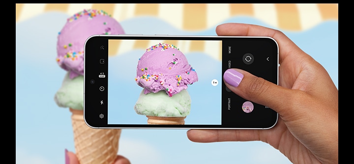 A hand taking a picture with a Galaxy S23 FE device seen from the front and horizontal. The screen in picture mode shows a clear and detailed picture of an up-close ice cream scoop with colorful sprinkles.