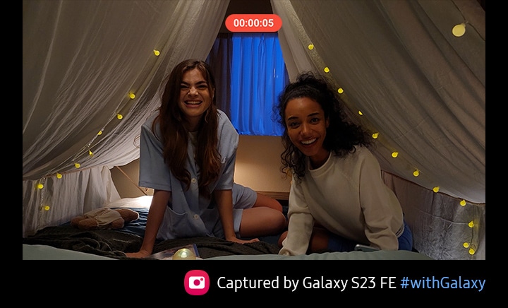 Two women smile and wave to come inside a beige cloth draped like a tent indoors as someone records a video of them. The room is dimly lit but the detail and color of the video is clear and balanced thanks to Nightography on the Galaxy S23 FE's rear camera. Text reads Captured by Galaxy S23 FE #withGalaxy.