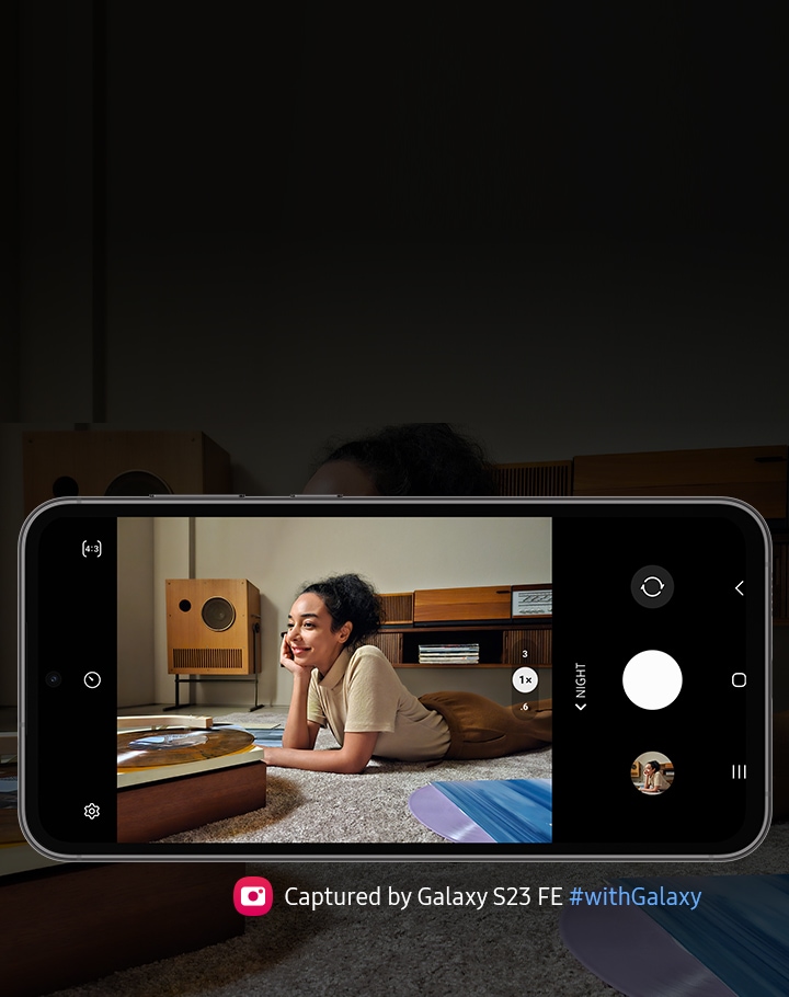 A Galaxy S23 FE device seen from the front and horizontal. The screen in picture mode shows a clear image of a woman listening to vinyl against the backdrop of a dimmer light setting. Text reads Captured by Galaxy S23 FE #withGalaxy.