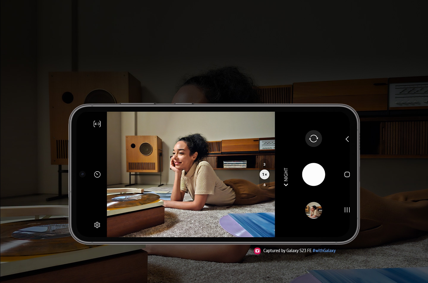 A Galaxy S23 FE device seen from the front and horizontal. The screen in picture mode shows a clear image of a woman listening to vinyl against the backdrop of a dimmer light setting. Text reads Captured by Galaxy S23 FE #withGalaxy.