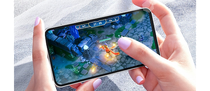 Two hands playing a video game on a Galaxy S23 FE device in bright daylight. The screen shows a clear image thanks to Vision Booster.