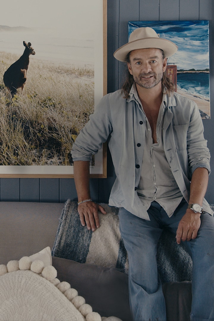 Interior stylist and designer Jason Grant in a blue-hued room at home. Jason posing in front of a gallery wall curated with coastal and nature-inspired pictures, designs and objects.