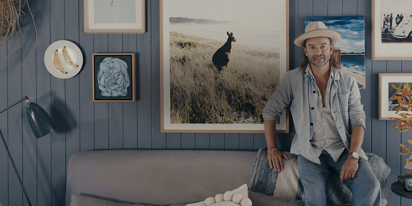 Interior stylist and designer Jason Grant in a blue-hued room at home. Jason posing in front of a gallery wall curated with coastal and nature-inspired pictures, designs and objects.