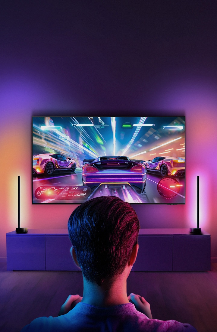 A person sitting in a dimly lit room playing a vibrant racing video game on a large-screen TV. The room is illuminated by colorful ambient lighting, enhancing the gaming experience.