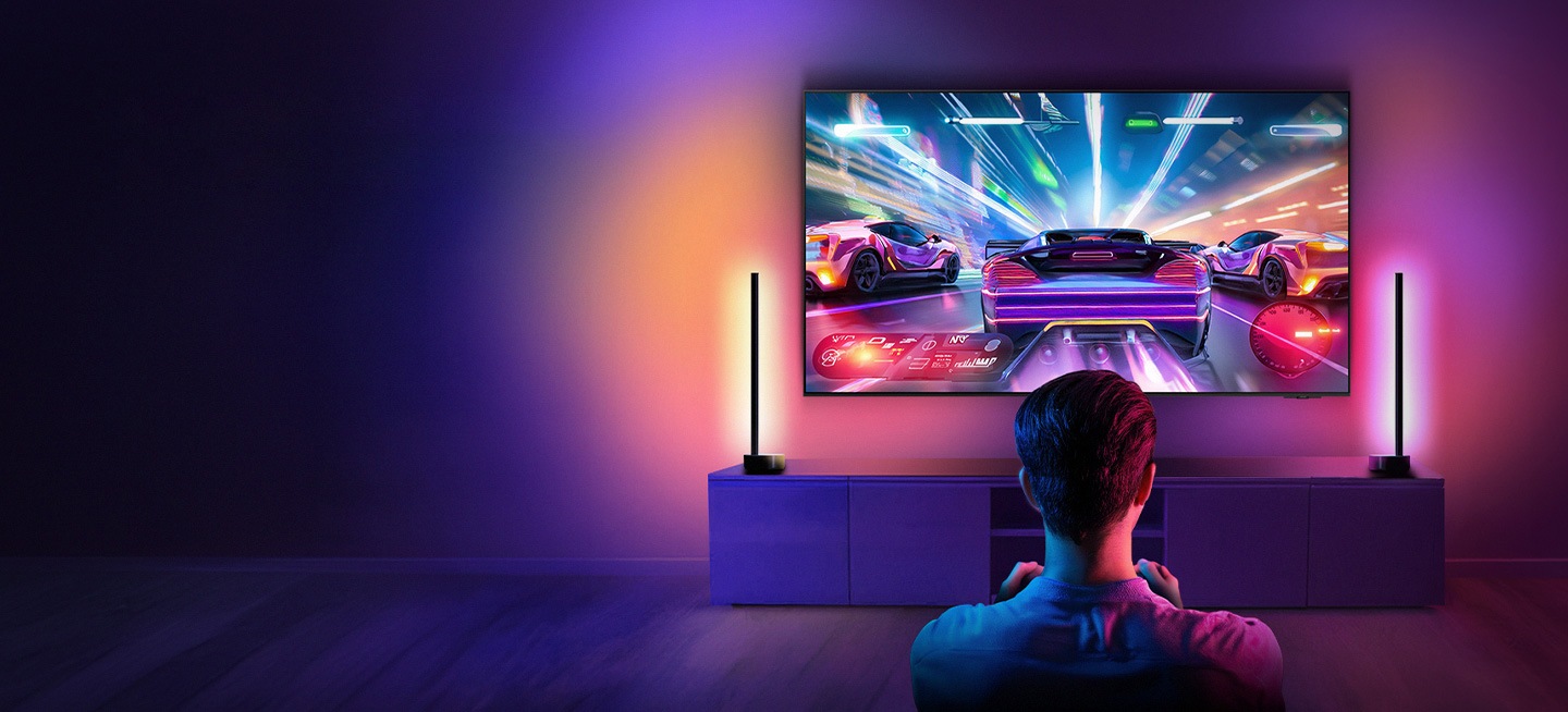 A person sitting in a dimly lit room playing a vibrant racing video game on a large-screen TV. The room is illuminated by colorful ambient lighting, enhancing the gaming experience.