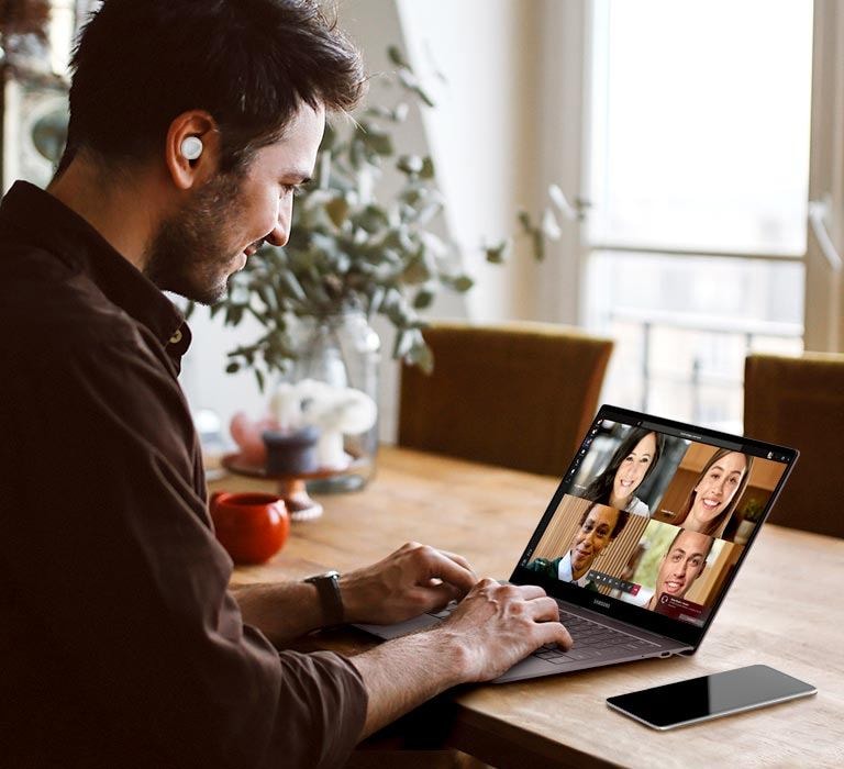 Man sits at home wearing white Galaxy Buds plus earbuds as he participates in a team conference call on his Windows PC.