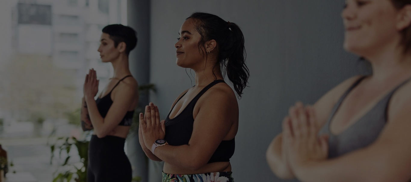 Three women in a yoga studio wearing workout clothes and standing with their hands in prayer position.