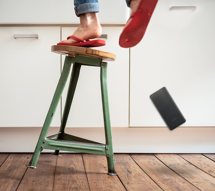 A closeup view shows a person's feet standing precariously on a wobbly kitchen stool. The stool is tilting to the right, with only one of its three legs on the floor, and the person's smartphone is falling to the ground in mid-air. 
