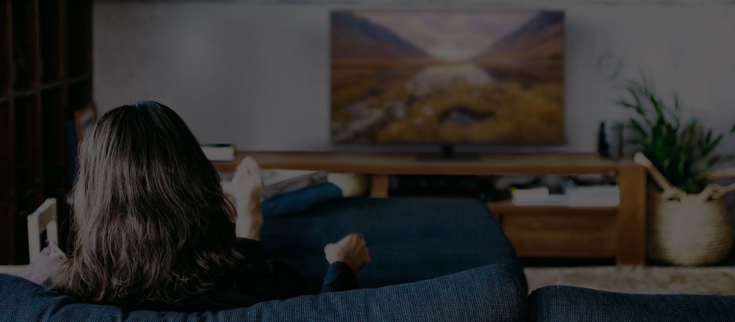Uma mulher, sentada num sofá na sala de estar, está a ver TV. 