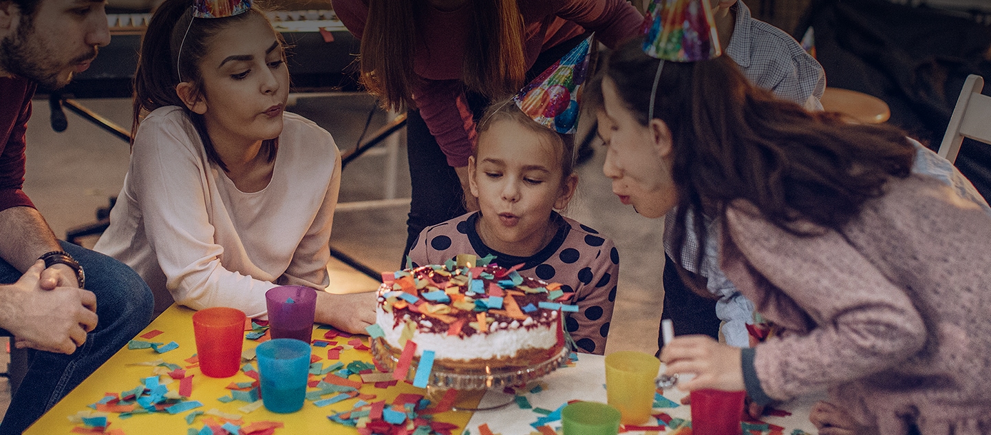 Uma festa de aniversário com crianças e adultos. Todos usam chapéus de festa. Rodeados por confettis, sopram as velas de um bolo que está no centro.