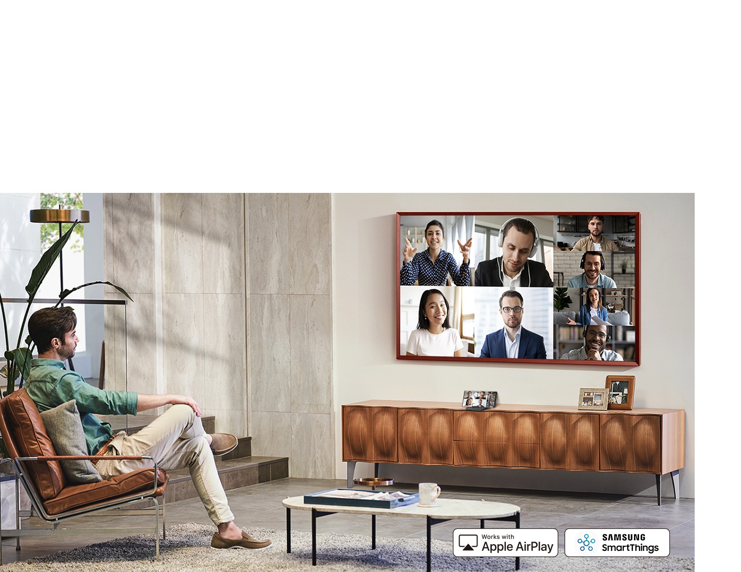 Un hombre está en un salón y utiliza la pantalla de su televisor para chatear por vídeo con varias personas. Debajo de su televisor está su smartphone que muestra el mismo videochat. Aparece el logotipo de Samsung SmartThings y Works with Apple AirPlay.