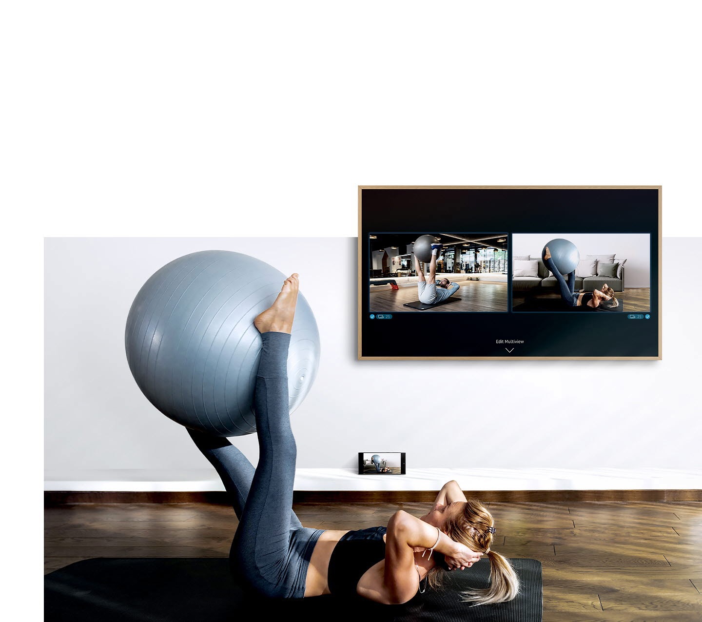 Una mujer se ejercita frente a su televisor. En la pantalla del televisor puede ver una pantalla de sí misma, así como la pantalla del entrenador físico, y debajo del televisor la cámara de su smartphone está capturando su entrenamiento para que pueda verlo en la pantalla del televisor.