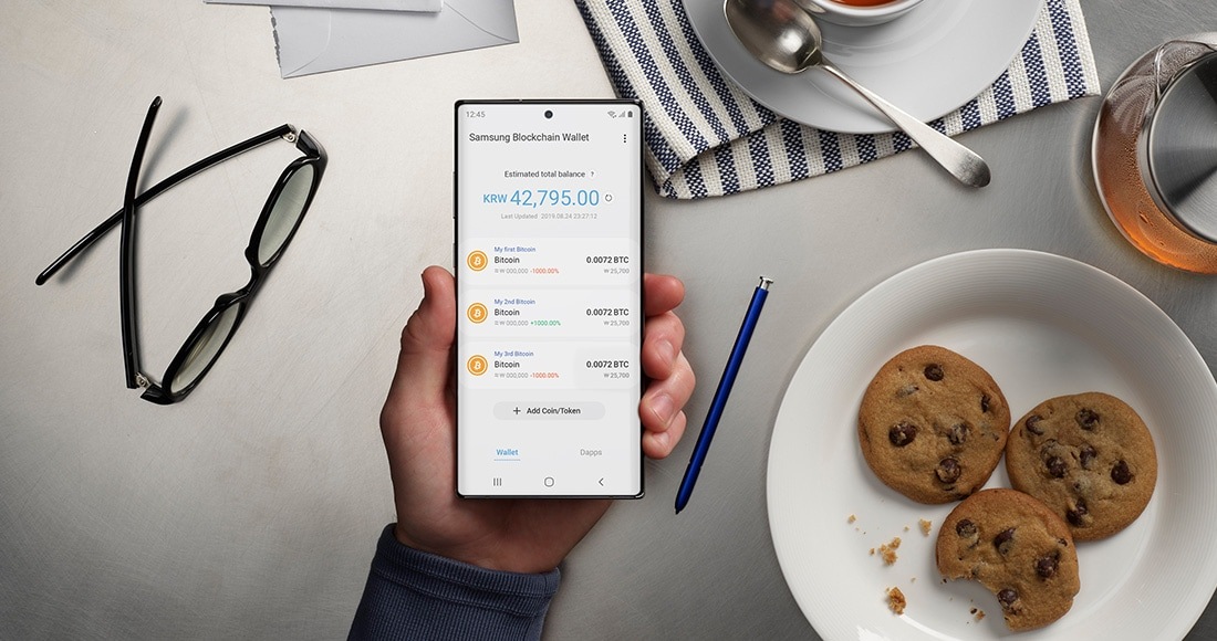Eine Hand hält das Galaxy Note10+ mit dem Samsung Blockchain Wallet Interface auf dem Display. Im Hintergrund sind Gläser, eine Platte mit Keksen und Tee zu sehen, die demonstrieren, wie einfach und sicher es ist, Blockchain-Transaktionen von zu Hause aus zu tätigen.