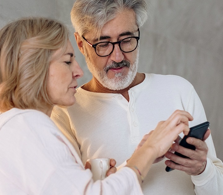 An older couple, both dressed in white. They are both looking at and inspecting something on the Galaxy smartphone the man is holding.