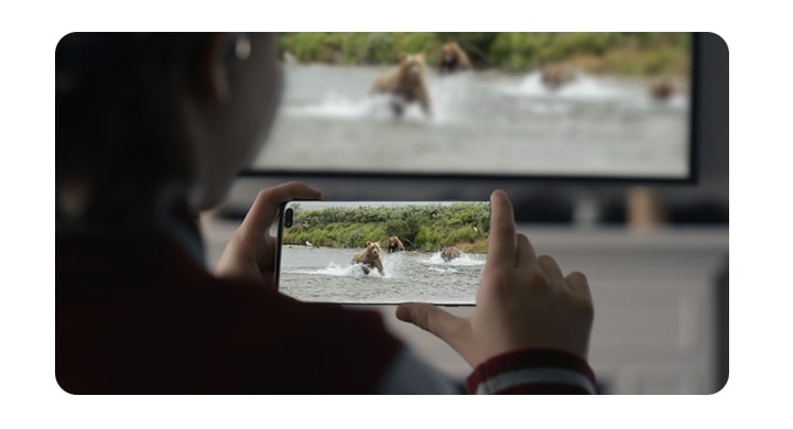 A young girl is holding a Galaxy Note10 in her both hands. And there is a video of bears in the water scene on her phone that is also on the TV in front of her