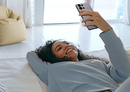 Une femme est allongée sur le sol, souriante, et regarde un Galaxy S25 Ultra, avec un pouf jaune visible derrière elle.
