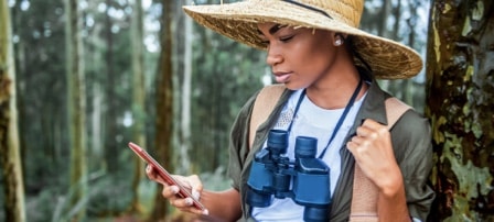 Uma mulher com um chapéu de abas largas e binóculos ao redor do pescoço verifica o celular enquanto se inclina contra uma árvore na floresta.